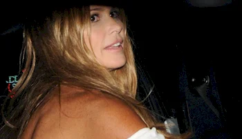 Close up of a woman with long, light brown hair looks over their shoulder, wearing a white off-the-shoulder top and smiling slightly. The photo is taken at night with partial shadow.