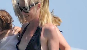 A woman with blonde hair in a ponytail, wearing sunglasses and a black swimsuit, smiles outdoors in bright sunlight.