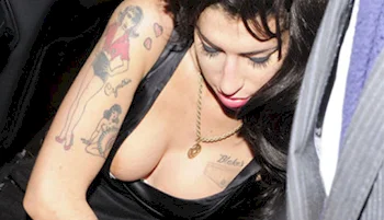 A woman with tattoos on her arm and chest, wearing a black top and gold necklace, looks downwards.