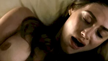 A close-up, high-angle shot of a woman with her eyes closed, lying down with her head on a pillow. Her mouth is slightly open, and she appears to be in a moment of intense intimacy. She has brown hair and is wearing a small earring.