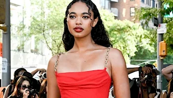 A woman with long curly hair wearing a red dress stands outdoors while photographers take her picture. City buildings and greenery are visible in the background.