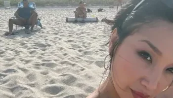 A woman takes a close-up selfie on a sandy beach with two people sitting on chairs in the background.