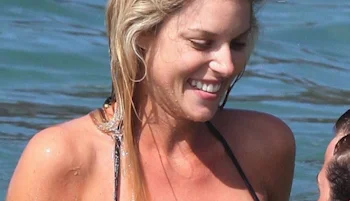 Woman with blonde hair in a black swimsuit smiles while standing in the water, with the ocean in the background.