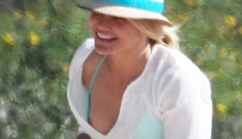 A woman wearing a blue swimsuit, white cover-up, and a sunhat smiles outdoors with blurred greenery and yellow flowers in the background.