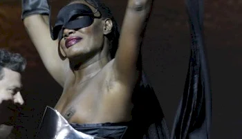 A black woman wearing a black mask and shiny metallic outfit raises their arms, smiling confidently on stage. The background is dark, highlighting their pose and expression.