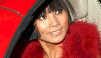 Asian woman with short dark hair and bangs, wearing a red fuzzy coat, smiles while sitting in a car with a red door.