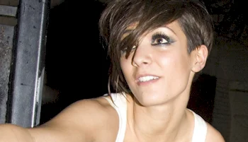 Woman with short, dark, side-swept hair and dramatic eye makeup, smiling and looking slightly upward; she is wearing a white sleeveless top.