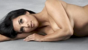 A professional studio portrait of a woman with dark hair, lying on her side on a light gray surface, looking directly at the camera.