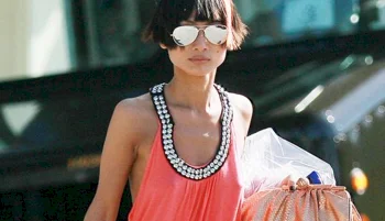 Asian woman with short hair and sunglasses wears a sleeveless, beaded-neck coral dress and carries a large handbag and shopping bag outdoors.