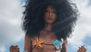 Woman with voluminous curly hair wears a bikini decorated with a starfish and a seashell, standing outdoors against a cloudy sky.