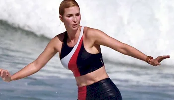 A woman with wet hair, wearing a sleeveless, color-block swimsuit, is balancing on a surfboard in the ocean with waves behind her.
