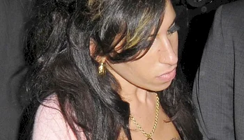 A woman with long dark hair, wearing gold jewelry and a light-colored top, looks down with a serious expression.