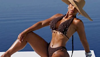A woman in a brown bikini and wide-brimmed hat poses on a white surface near calm blue water, with one leg bent and arm resting on her knee.