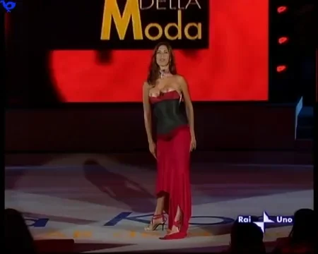 VeronicaMayaDoubleNipSlip2 A woman in a strapless red dress and black corset stands on a stage with Moda displayed behind her. The stage is lit, and logos are visible at the bottom right corner of the image.