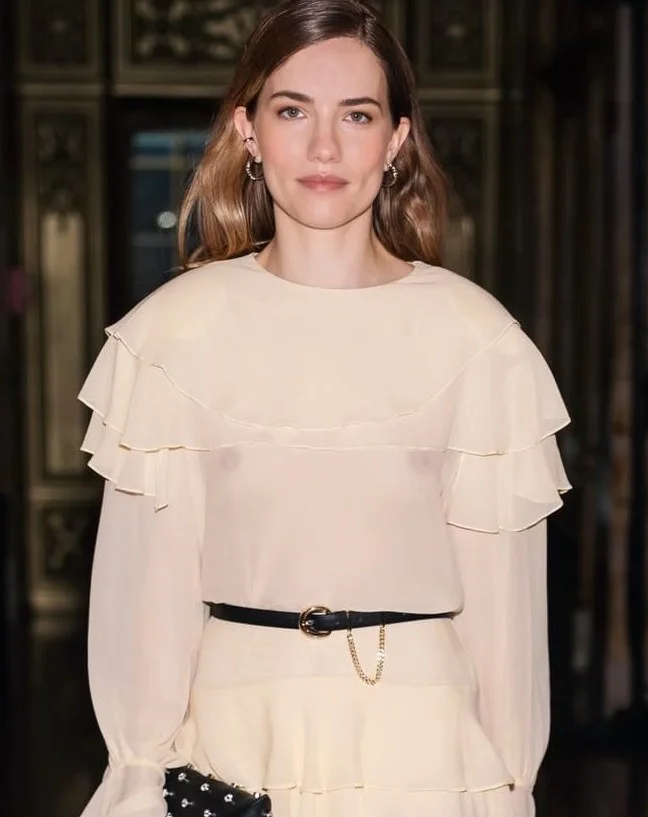 WillaFitzgeraldInSheerDressInstaPic1 A woman with long brown hair wears a cream ruffled blouse and matching skirt, accessorized with a black belt and small hoop earrings, standing indoors with ornate dark background details.