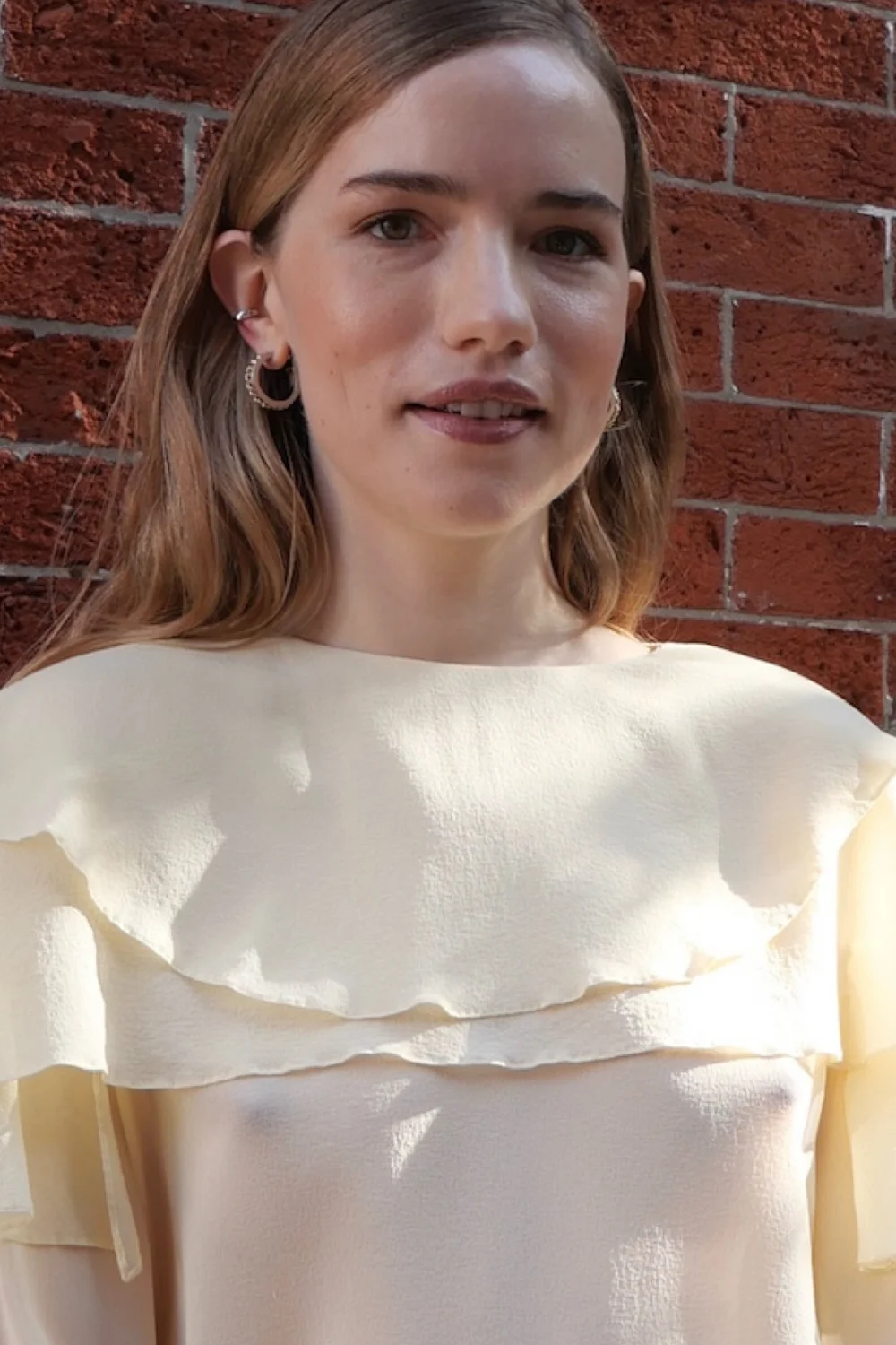 WillaFitzgeraldInSheerDressInstaPic2 A woman with long hair stands in front of a red brick wall, wearing a cream-colored top with ruffled layers and hoop earrings. She is looking slightly past the camera.