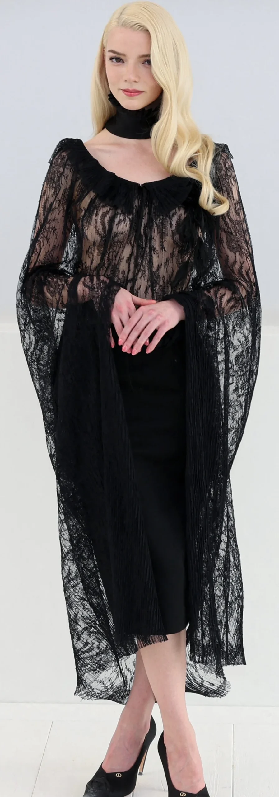 A blonde woman wearing a black, sheer lace gown with wide sleeves and a black choker.
