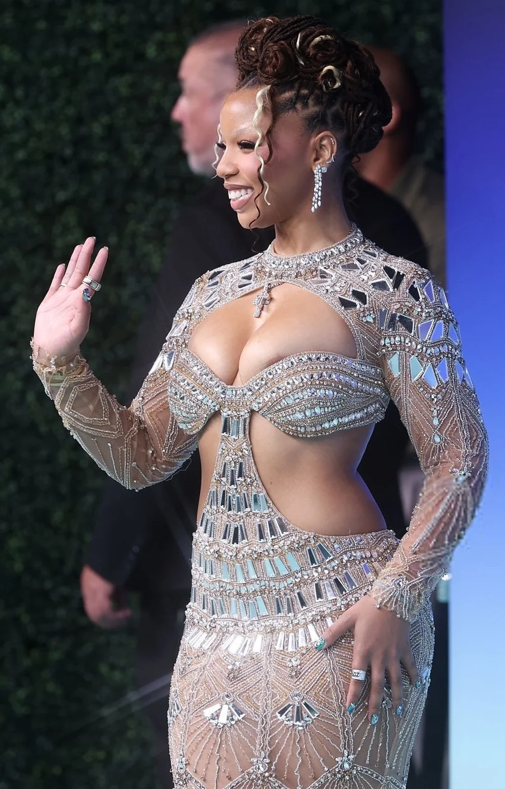 ChloeBailey2026MTVVMAPic2 Chloe Bailey waves while wearing a floor-length, long-sleeved gown embellished with intricate silver beadwork and mirrored glass panels.