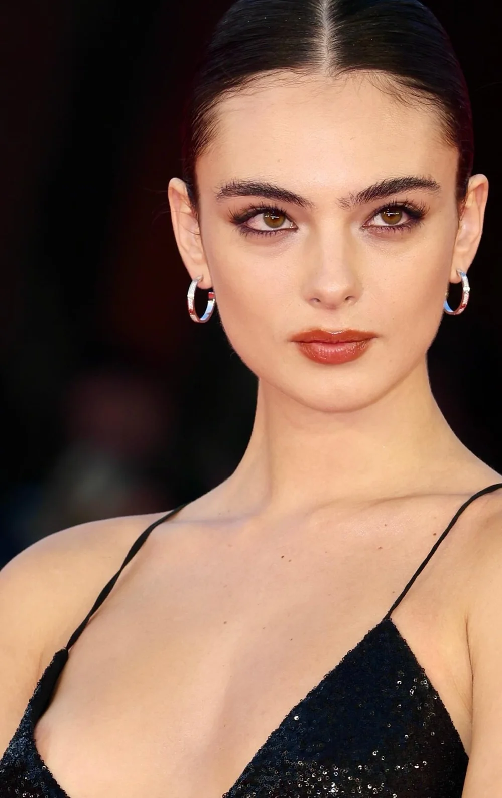 DevaCasselNipSlipLeFantomeDeLOperaRomeFFPic2 A close-up portrait of a woman with dark hair pulled back, wearing small silver hoop earrings and a black dress.