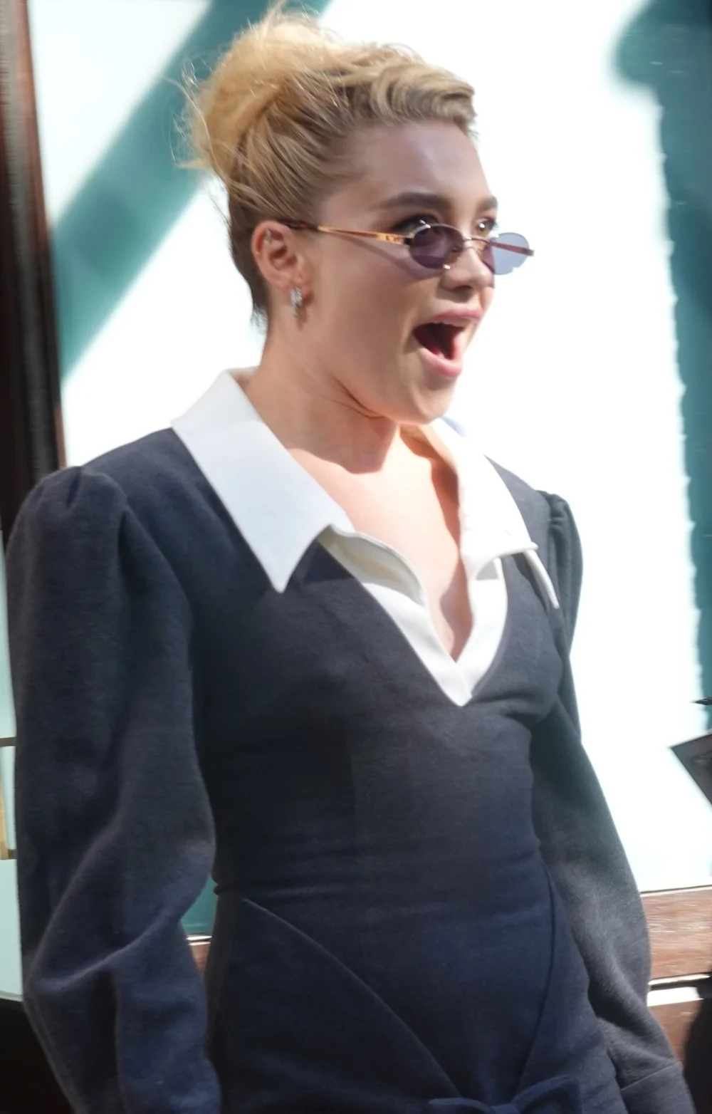 Florence Pugh wearing sunglasses and a black dress with a white collar, looking surprised with her mouth open.