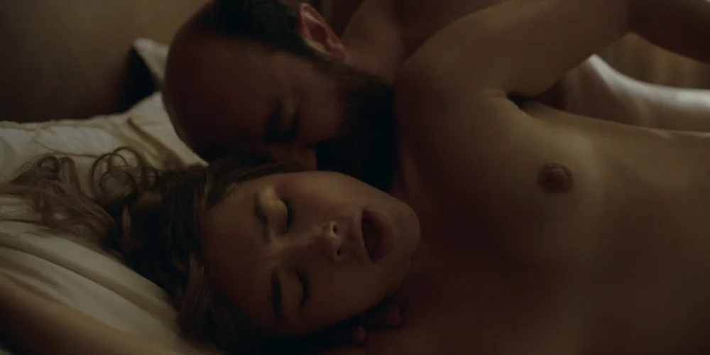 FlorencePughToplessEastOfEden1 Close up of topless Florence Pugh lying on her side in a dimly lit room. A man is behind her. They are having sex.