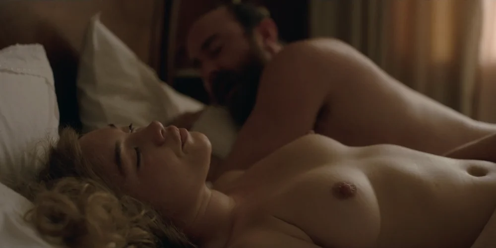 FlorencePughToplessEastOfEden3 A topless Florence Pugh lying on her back in a dimly lit room next to a man. He turns his head and looks at her.