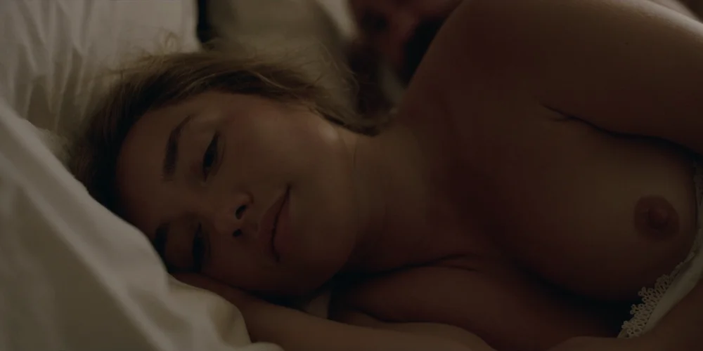 FlorencePughToplessEastOfEden4 Close up of topless Florence Pugh lying on her side in a dimly lit room. She has a slight smile on her face.
