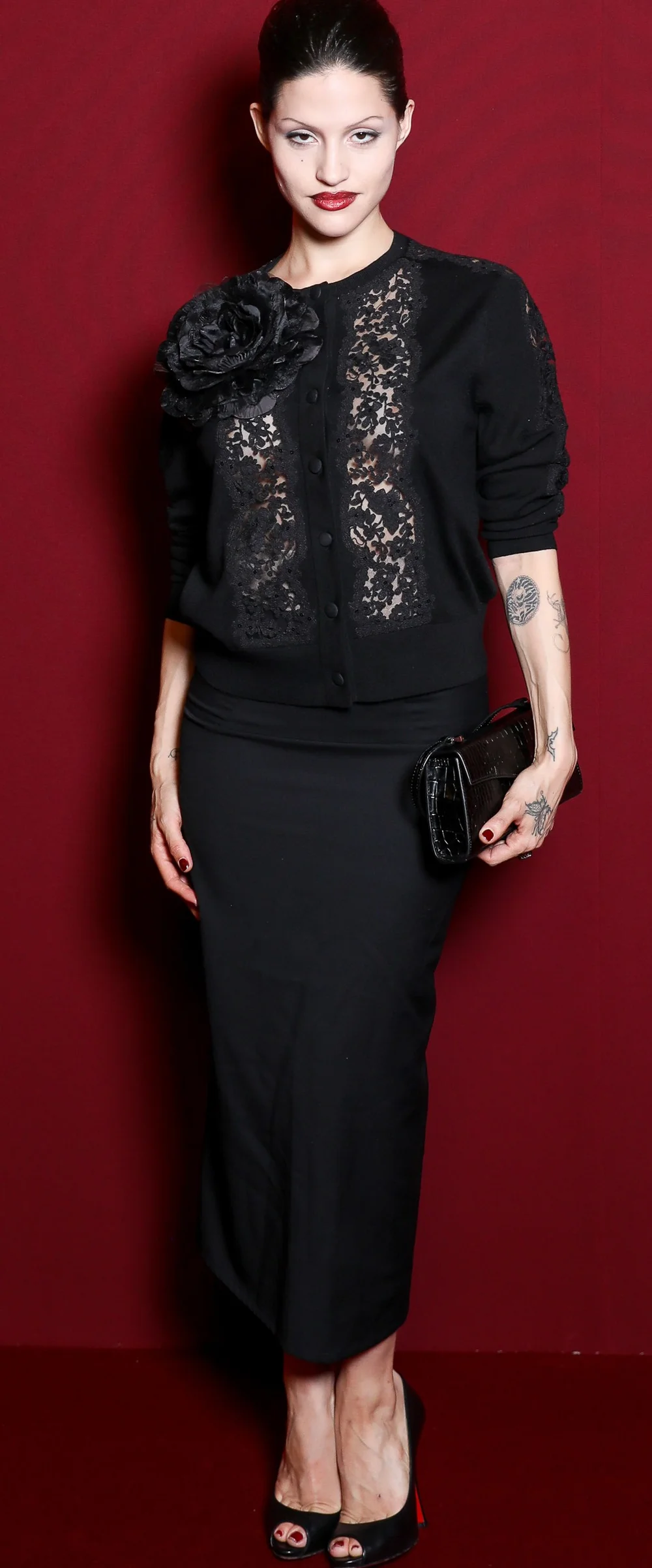 GabbrietteH&MElieSaabParisPic1 A woman wearing a black cardigan with a floral embellishment and lace panels over a matching long skirt, holding a black clutch.