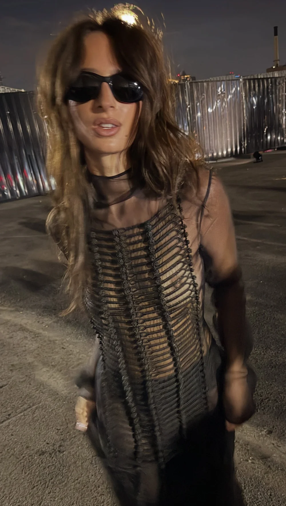 A woman with long brown hair wearing dark sunglasses and a sheer black top with a vertical rope-like corded design.
