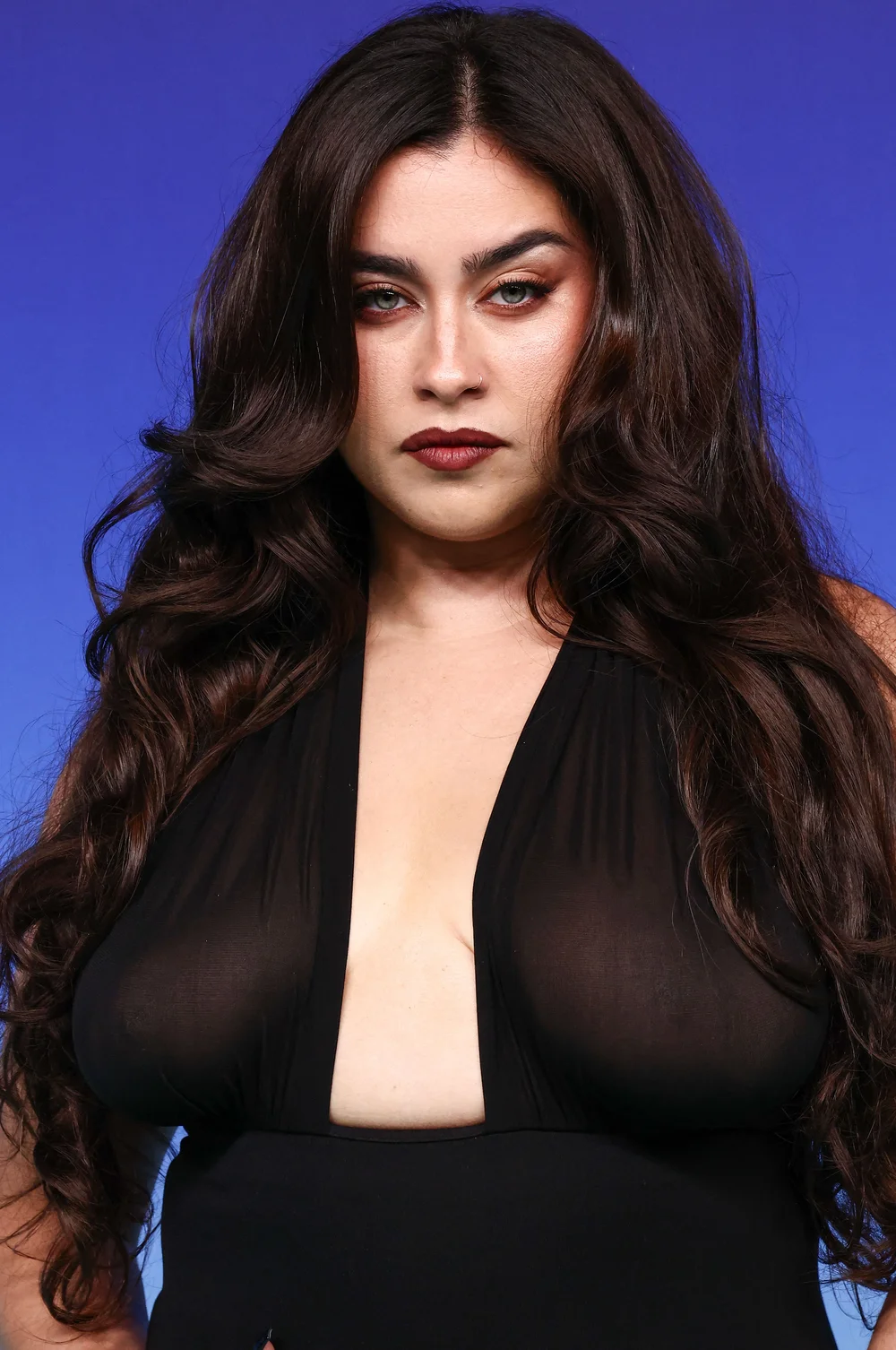 LaurenJauregui2026MTVVideoMusicAwardsPic1 A woman with long, wavy dark hair wears a black top with a deep plunging neckline against a blue background.