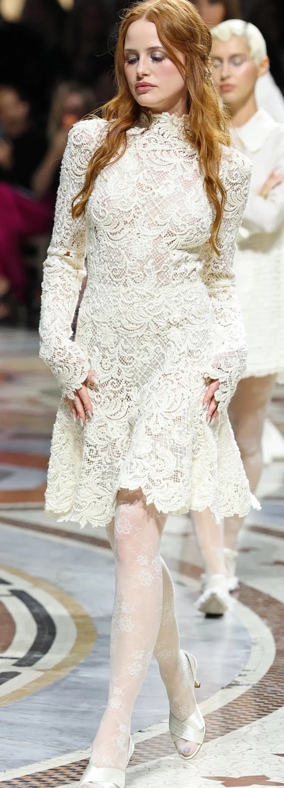 MadelainePetschNipsRunway26VogueWorldPic1 A model walks a runway wearing a long-sleeved cream lace mini dress and matching lace tights.