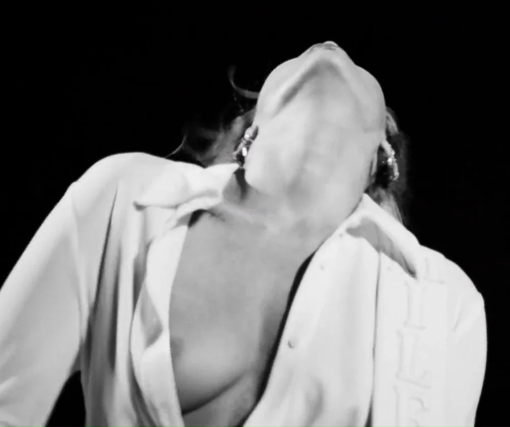 MileyCyrusNipSlipDifferentReligion Miley Cyrus in an unbuttoned white shirt tilts their head back against a black background.