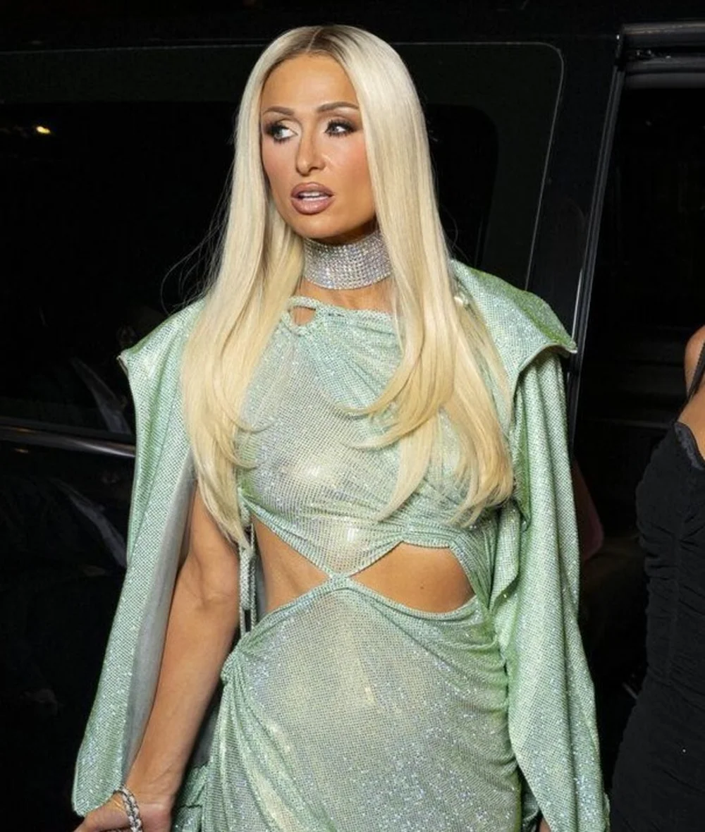 ParisHiltonSeeThrough26PFWPic2 Paris Hilton with long blonde hair, wearing a sparkly light green cutout dress and a crystal choker.