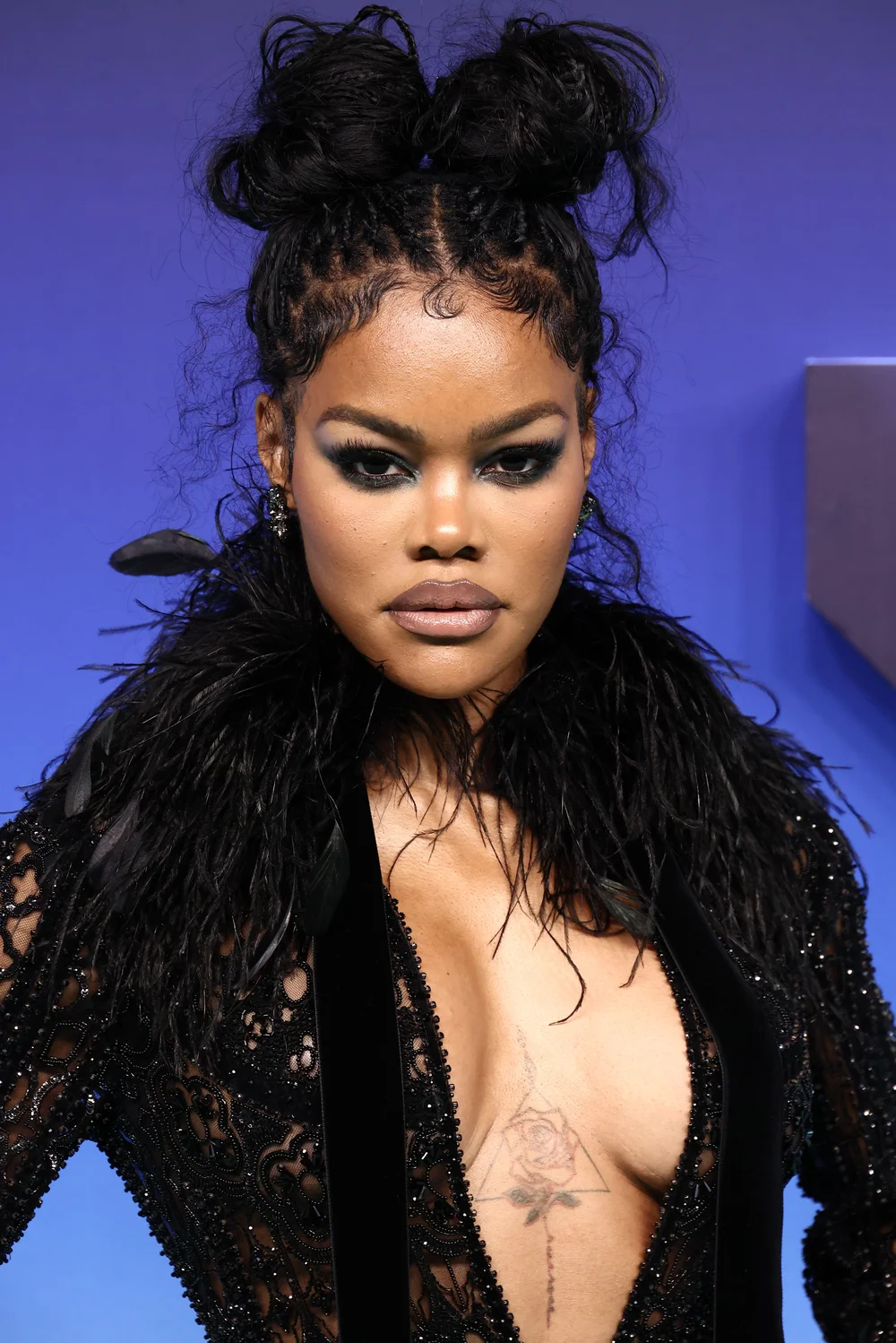 TeyanaTaylor2026MTVVideoMusicAwardsPic1 Portrait of Teyana Taylor wearing a black feathered garment with a geometric chest tattoo against a blue background.