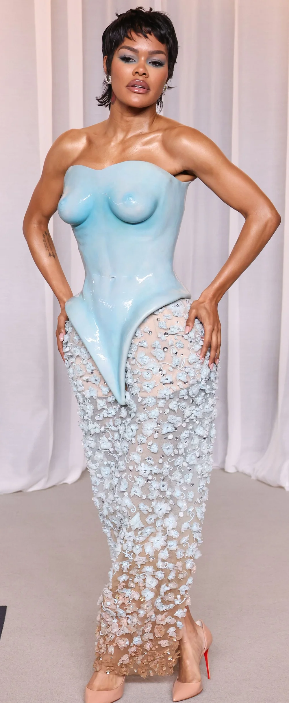 TeyanaTaylor6thAcademyMuseumGalaPic2 A woman poses wearing a structured, light blue, molded bodice shaped like a torso over a sheer, floor-length skirt covered in light blue floral appliqués.