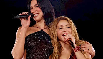 Two women stand close together while singing into microphones.