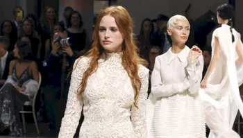Models walk a runway in white lace and structured dresses during a fashion show.