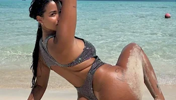 A woman in a sparkly cutout swimsuit posing on a sandy beach by the ocean.