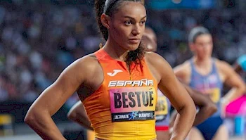 A female sprinter wearing an orange and yellow Spain jersey with her name, Bestué, printed on the chest, stands on a track during a race.
