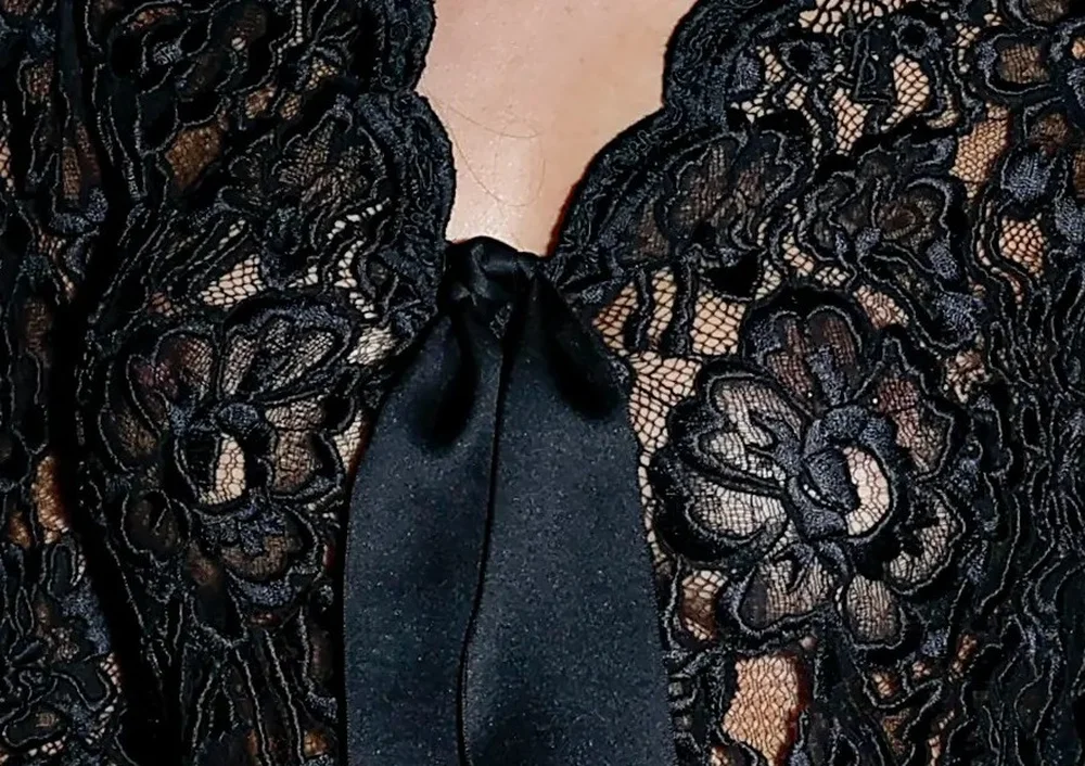 ZoeKravitzSeeThroughSLShow26PFWPic2 A black lace garment featuring floral patterns and a central ribbon tie at the neckline.